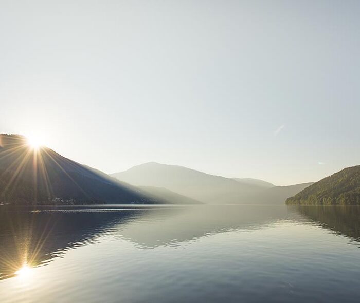 Der Millstätter See zwischen Berge und Hügel. Im Hintergrund geht die Sonne auf.