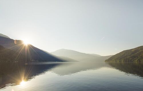 Der Millstätter See zwischen Berge und Hügel. Im Hintergrund geht die Sonne auf.